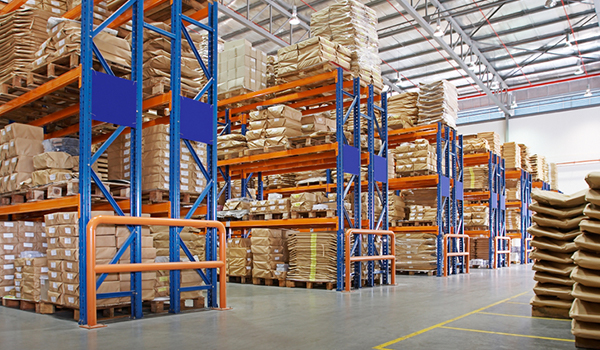 A spacious warehouse filled with stacked boxes on blue and orange shelving units situated beneath a high ceiling.