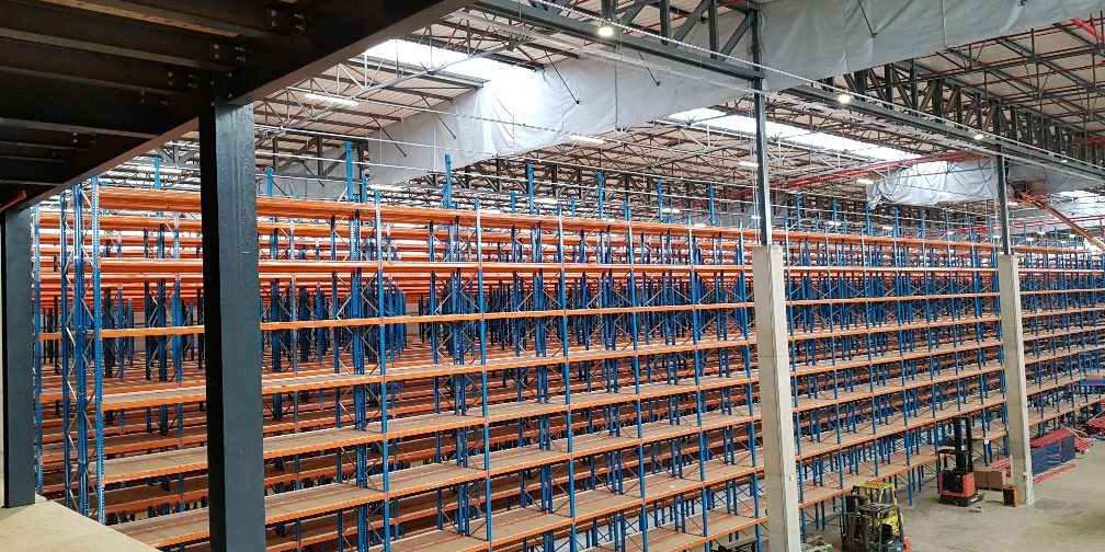 A blue and orange steel and wooden industrial shelving structure sitting high in a large empty warehouse next to a forklift