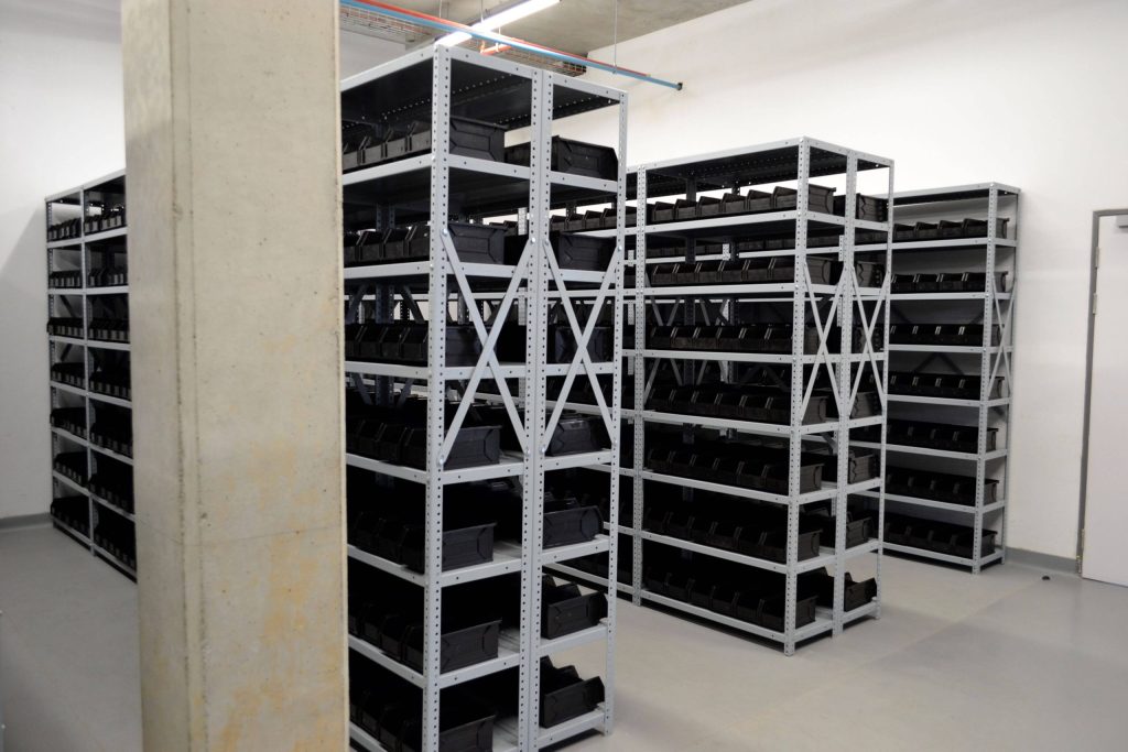 White and well-lit storage room with assembled steel bolted shelving lined with black organisers, comparing mobile shelving vs. bolted shelving