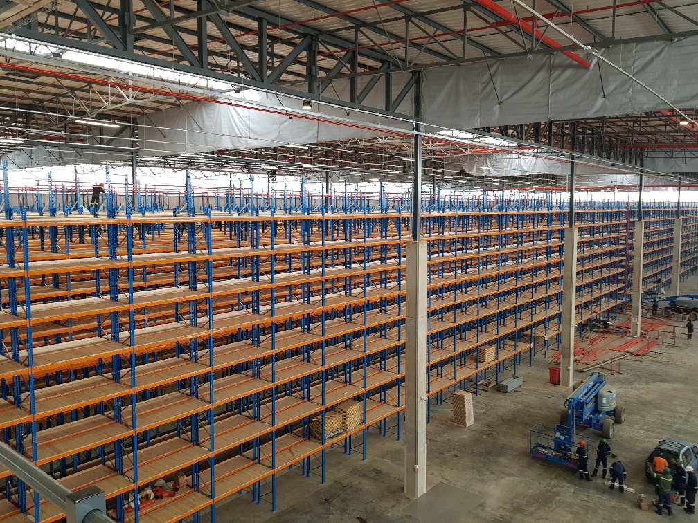 A tall blue and orange warehouse shelving structure in a large warehouse with workers beside it