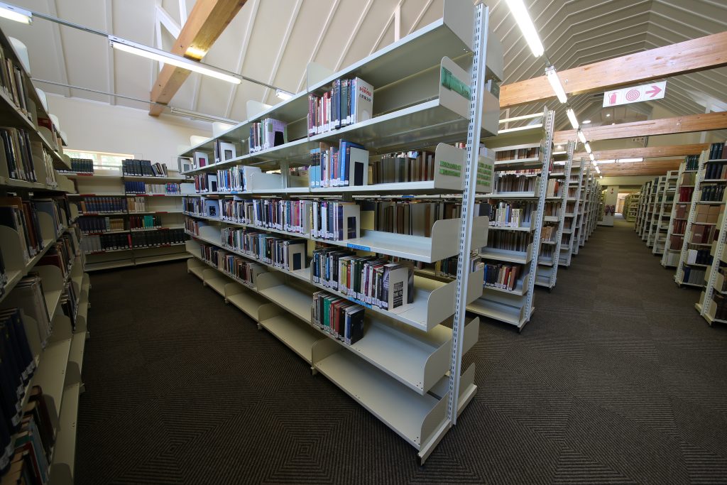 Library shelving with books, carpeted floor, and wooden beams on the ceiling.