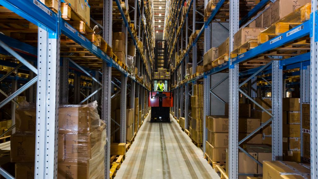 Warehouse aisle with tall shelves filled with cardboard boxes and a forklift operator in a high-visibility jacket.