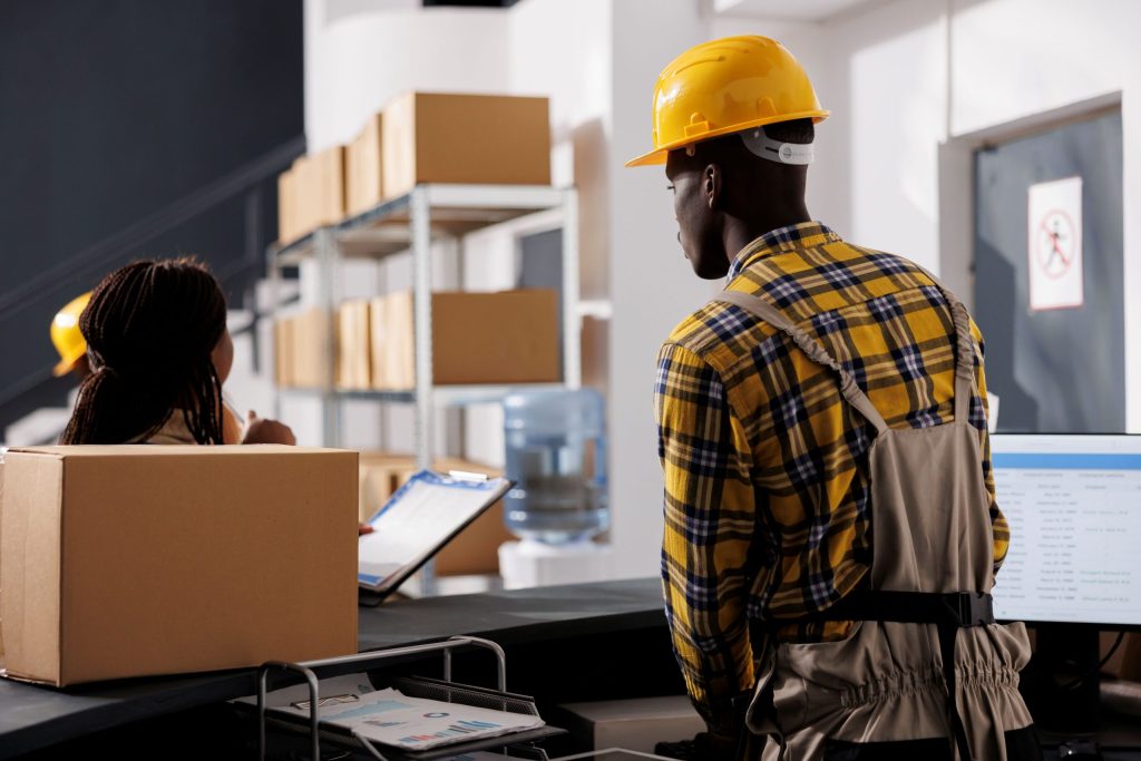 A worker in a plaid shirt and hard hat observes a colleague at a desk, surrounded by cardboard boxes and a water cooler.