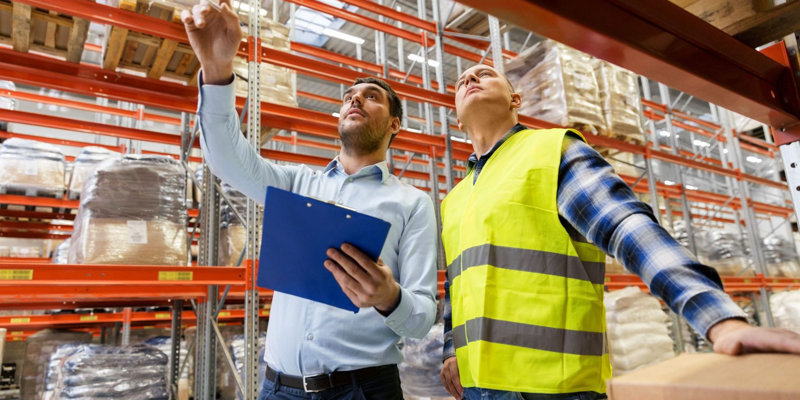Drive-Through vs. Pallet Shuttle Racking, Pros & Cons A worker and a businessman with a clipboard at a warehouse examining drive-through vs. pallet-shuttle racking