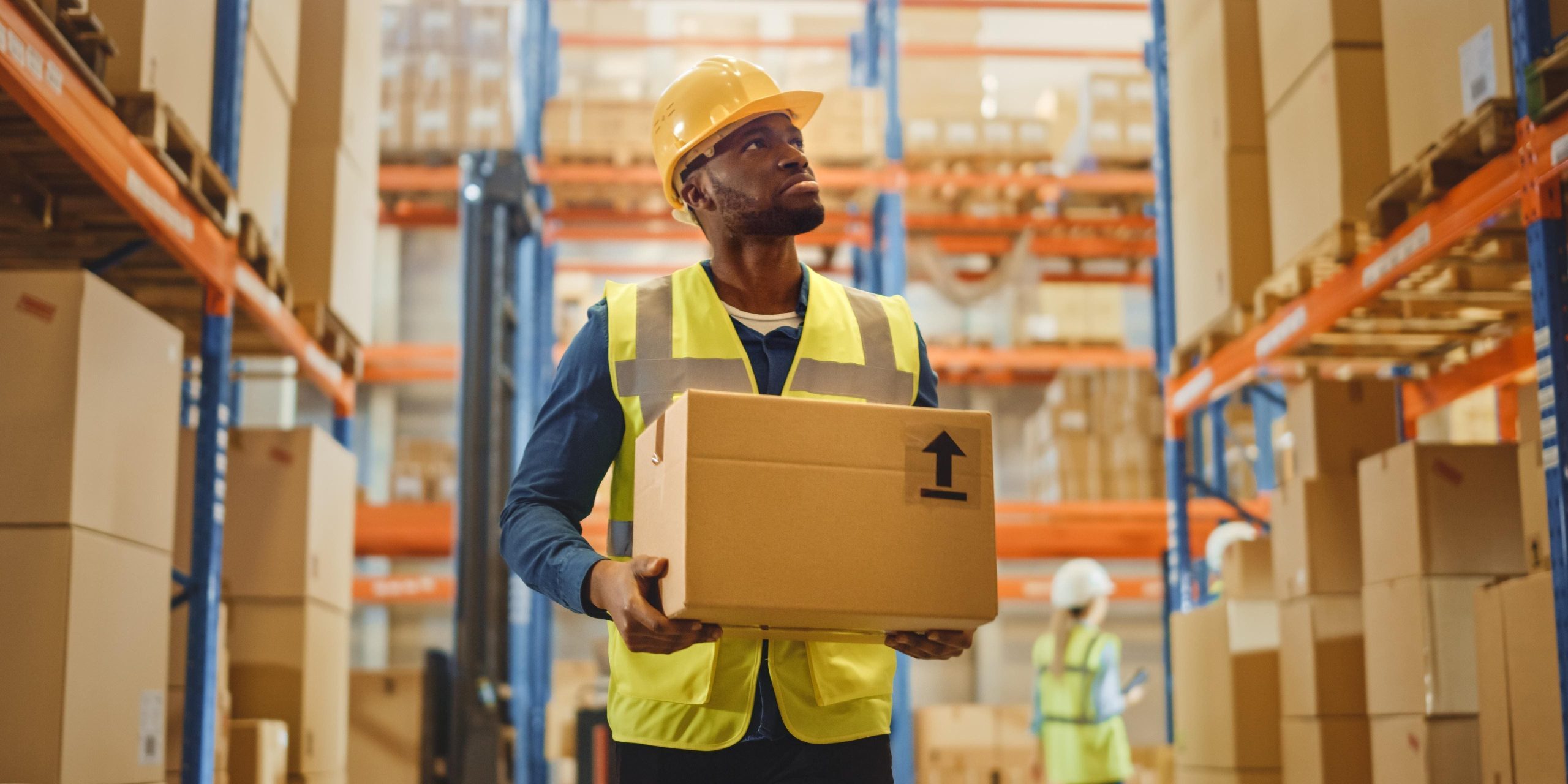 man-hard-hat-holds-box-warehouse (1) A worker carrying a box in a warehouse using an efficient warehouse dispatch process