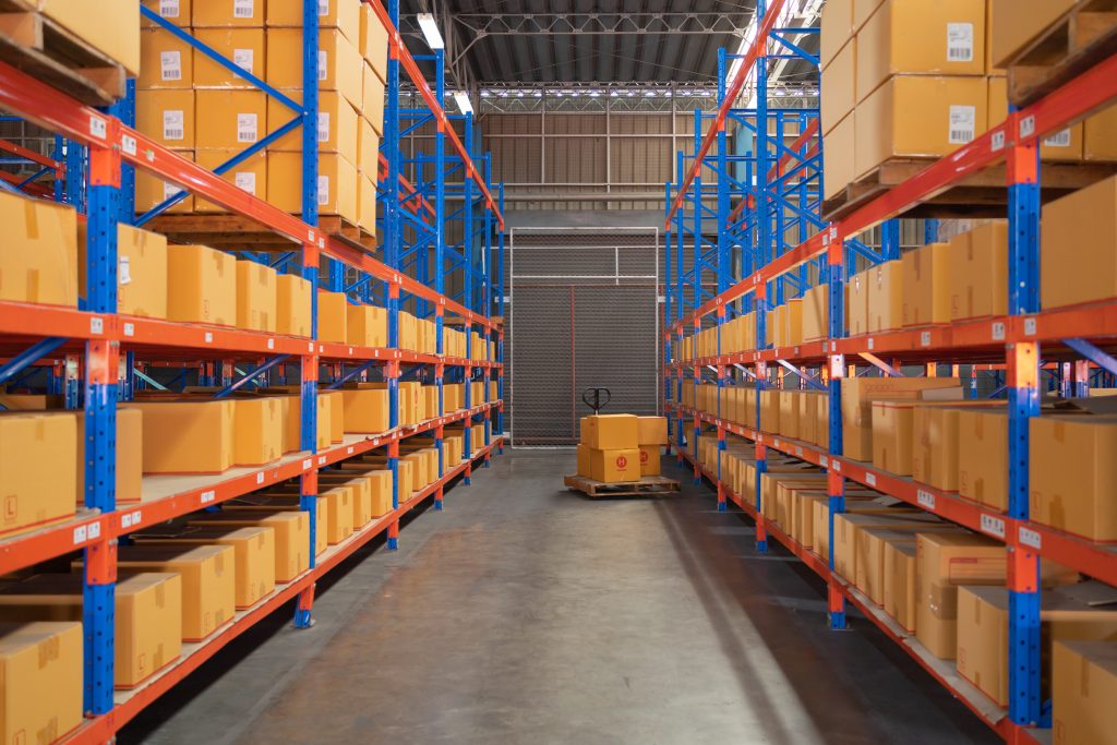 Warehouse featuring organized shelving units with orange and blue racks, stacked with boxes, and a pallet truck in the aisle.