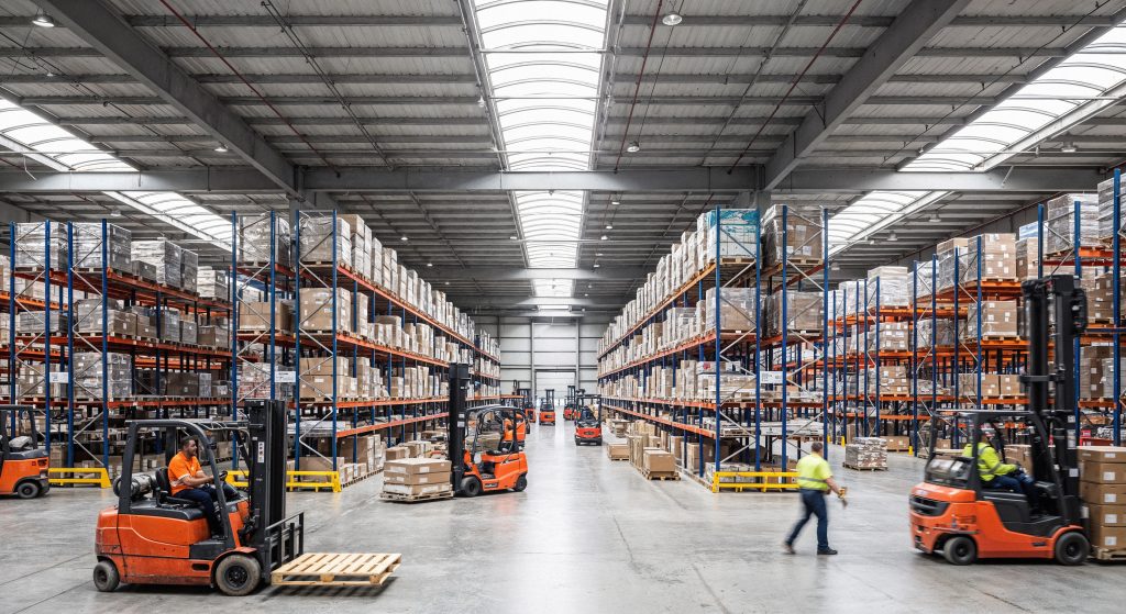 Busy warehouse interior featuring large shelving units filled with boxes, while forklifts and workers move goods throughout the space.