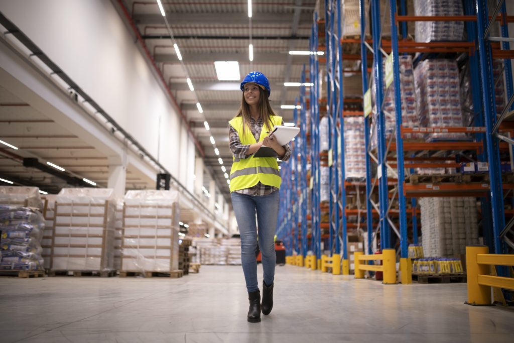Worker in safety gear walks through warehouse aisle, checking inventory on shelves with a clipboard, highlighting ergonomic shelving setup.