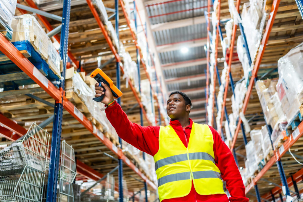 Warehouse worker in a hi-vis vest using a barcode scanner on palletised stock stored in tall pallet racking in a logistics and distribution centre