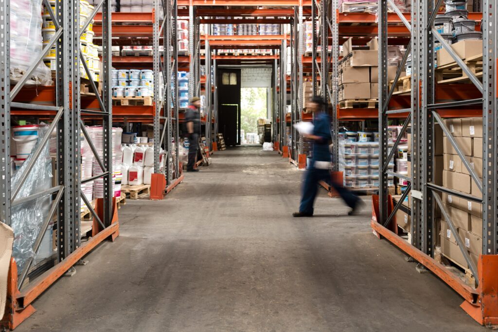 Retail racking and shelving system in a busy warehouse, holding pallets of paint buckets and boxed stock.